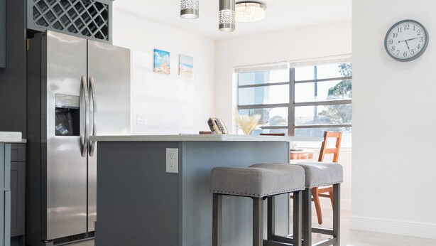 Una cocina moderna con un refrigerador de acero inoxidable, una isla gris con taburetes de bar y un amplio ventanal que proporciona luz natural.