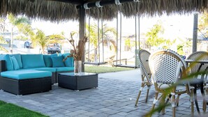Un espacio al aire libre con techo de paja, mobiliario de mimbre y vistas a palmeras y un vehículo al fondo.