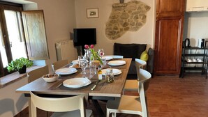 Een eetgedeelte met een houten tafel voor vier personen, witte stoelen, een stenen muur, een televisie en een keuken met een koffieapparaat.