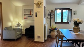 Ett vardagsrum med en soffa, ett matbord med stolar, en vas med blommor, en klädhängare och ett fönster med utsikt över grönområden.