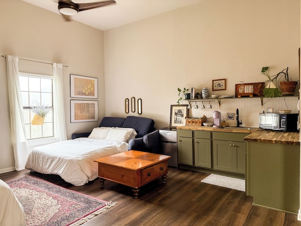 A cozy room with a bed, a wooden coffee table, a sofa, a kitchen area with cabinets and a microwave, and a window with curtains.