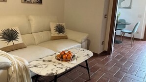 Een woonkamer met een bank, een bijzettafeltje met een schaal oranje en een eethoek met een tafel en stoelen.