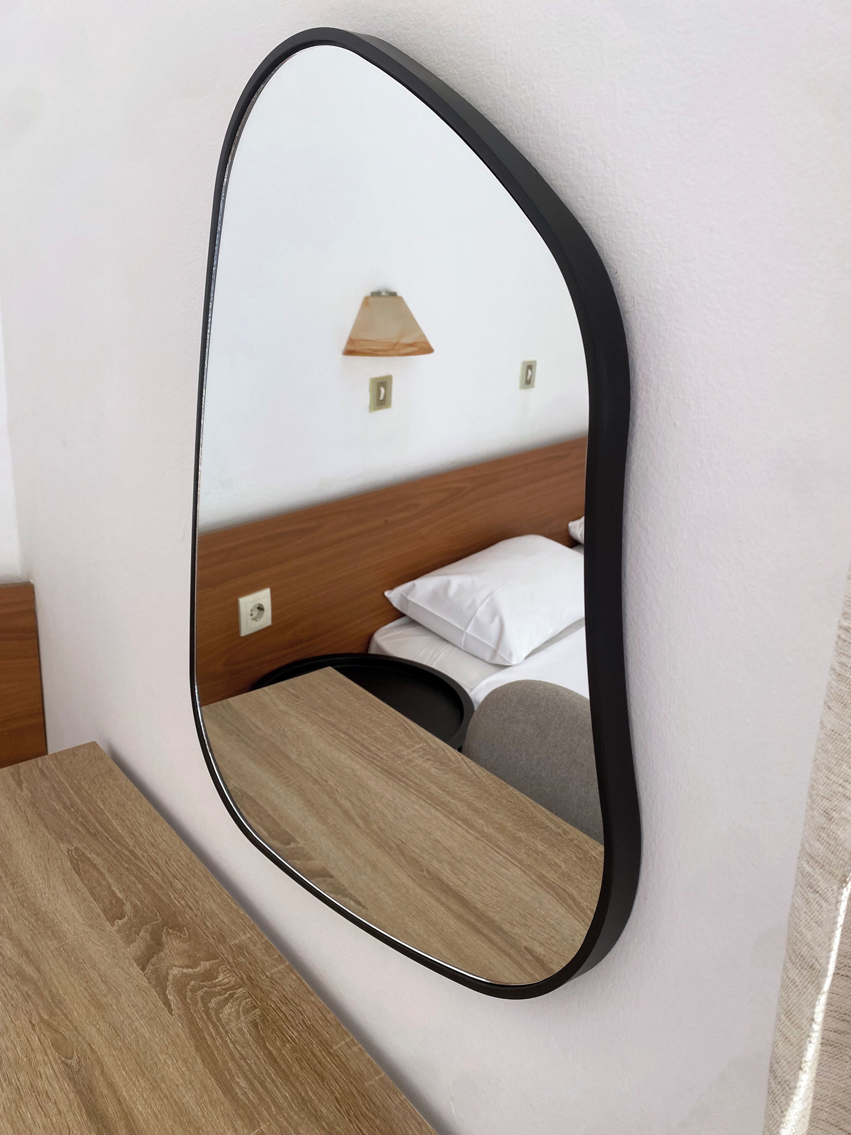 Un miroir avec un cadre en bois qui reflète un lit et une lampe fixée au mur.