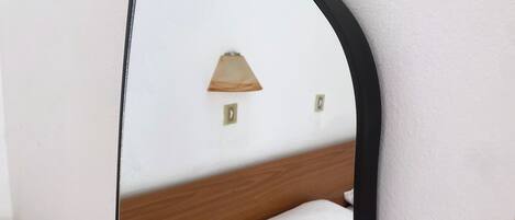 A mirror with a wooden frame reflecting a bed and a wall-mounted lamp.
