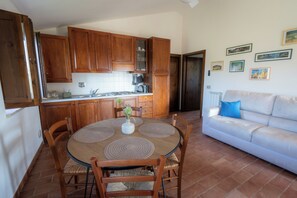 Cucina con mobili in legno, tavolo da pranzo con sottopiatti, divano bianco e pavimento piastrellato.