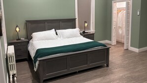Quarto com cama de madeira escura, roupa de cama branca, manta verde, mesa de cabeceira com abajur e radiador.