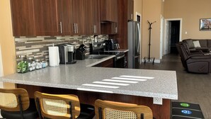 Cozinha moderna com armários de madeira escura, bancada de granito e geladeira de aço inoxidável. Há duas banquetas com assentos trançados e almofadas pretas.