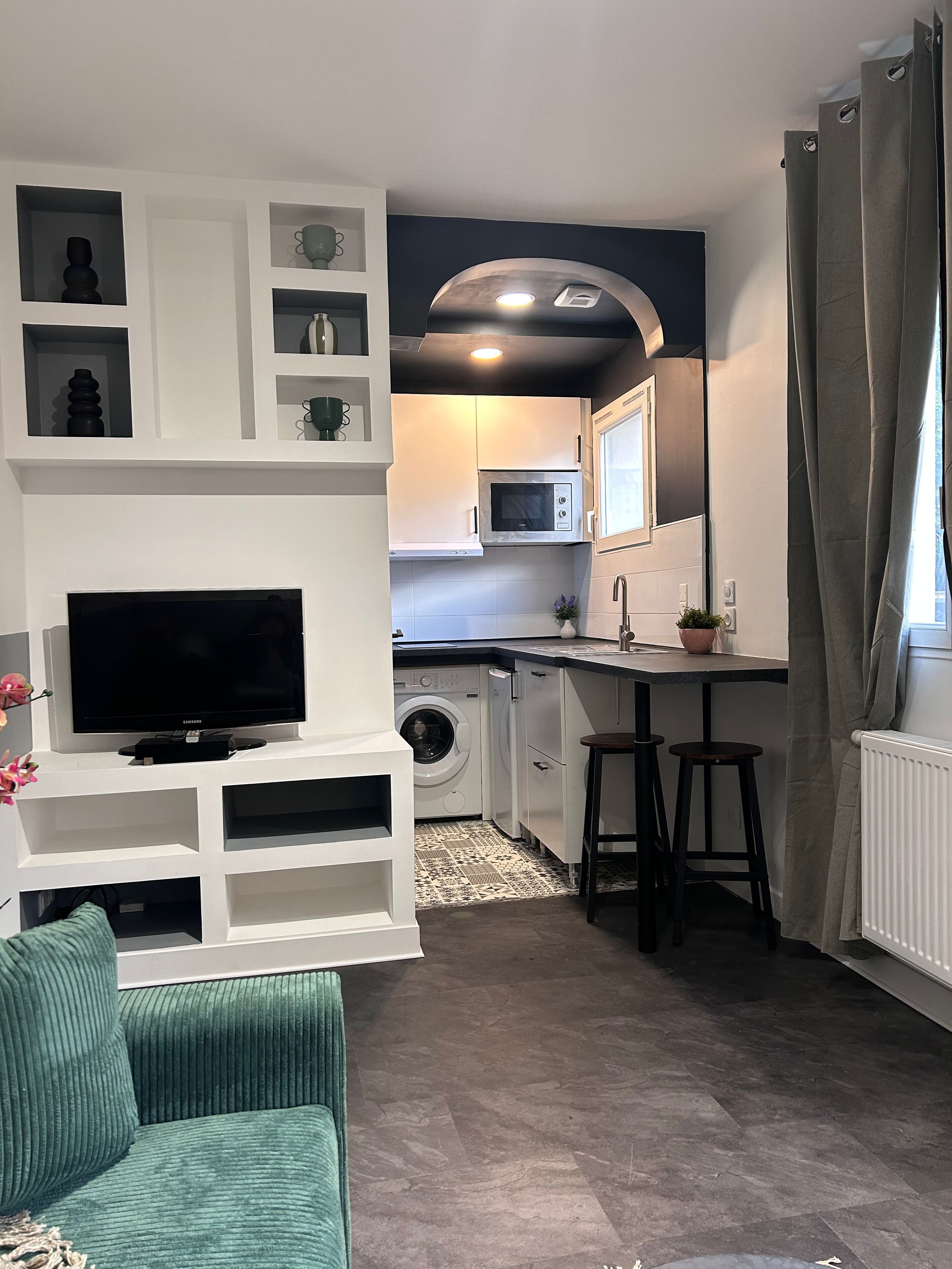 A compact living space with a flat-screen TV, a sofa, a kitchen area with a washing machine, and a dining area with stools.