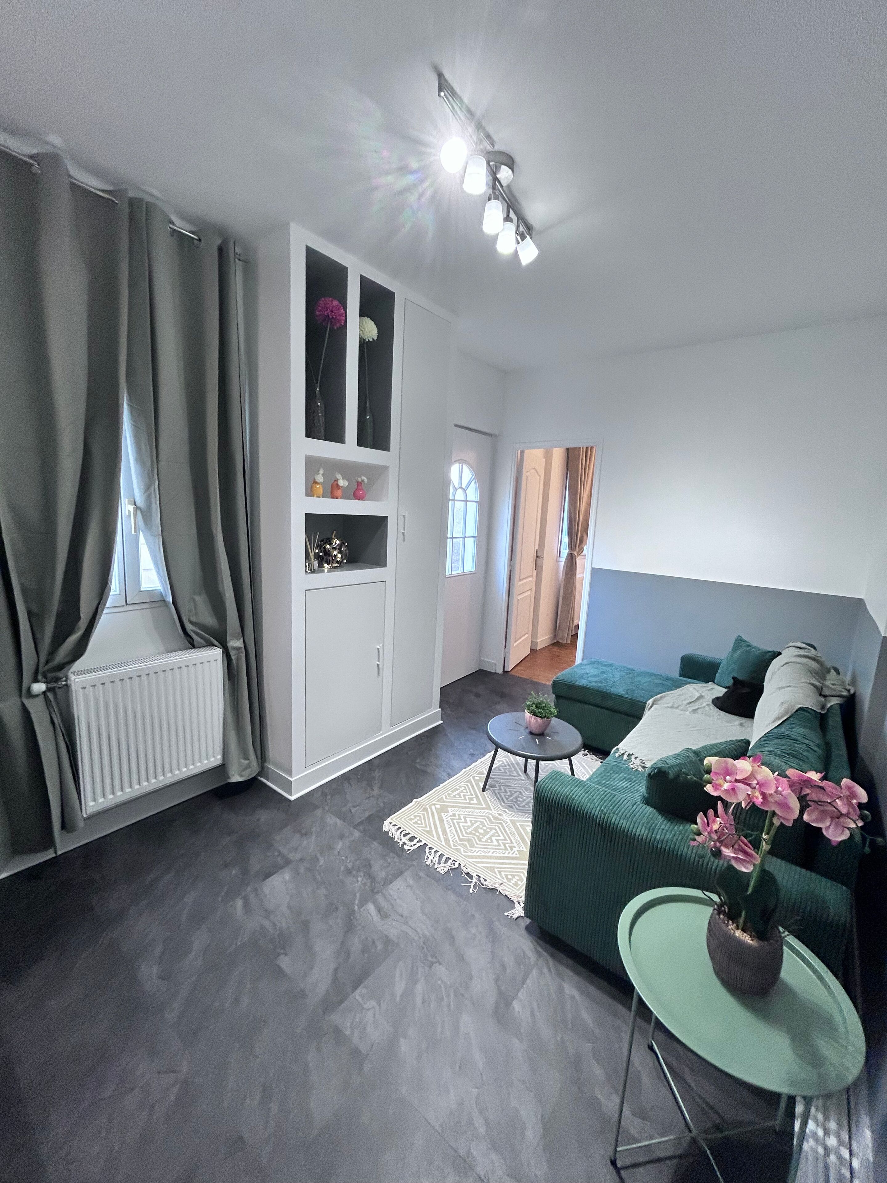 A modern living room with a grey sofa, a small round table, and a radiator. There is a white cabinet with glass doors and a plant on top.