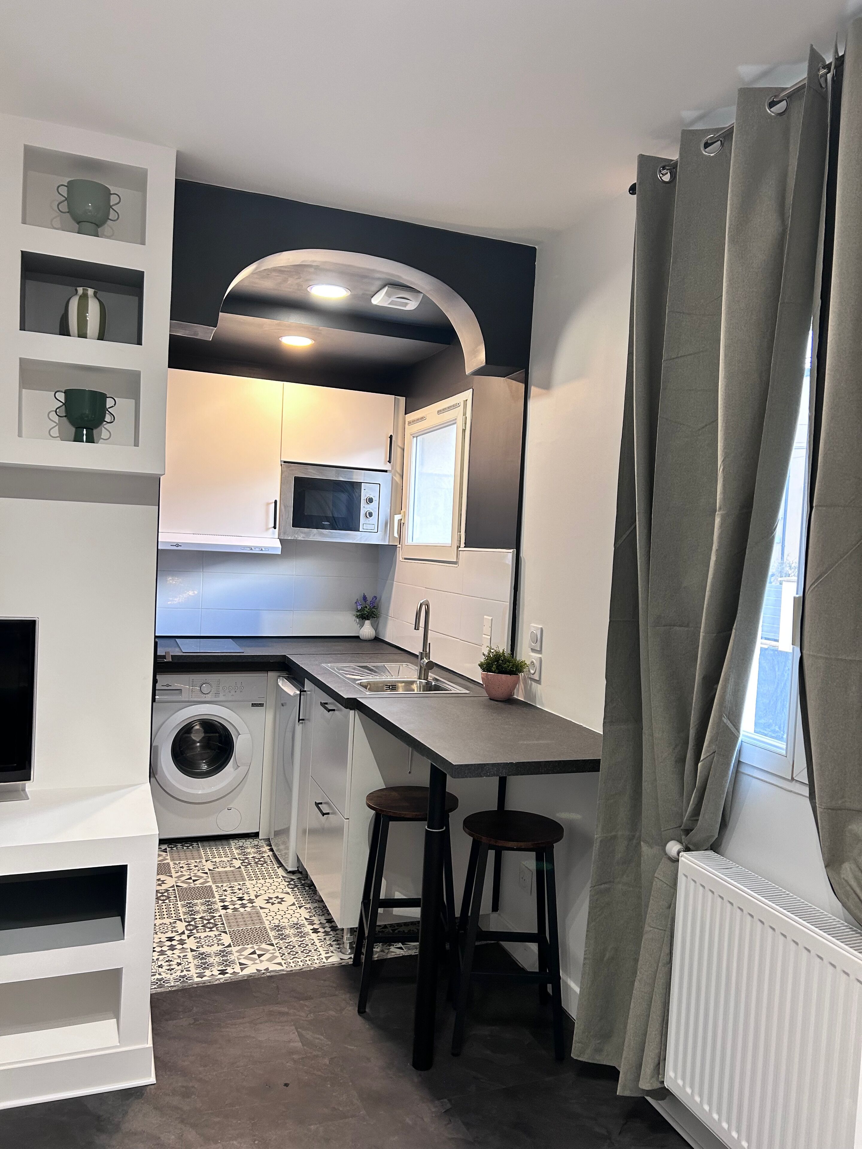 Cocina compacta con lavadora integrada, microondas y lavabo. Hay dos taburetes y una planta en maceta sobre la encimera.