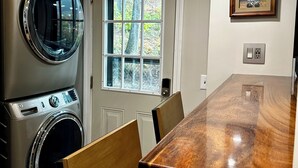 Ein Waschraum mit einer Waschmaschine und einem Trockner, ein Holzesstisch mit Stühlen, ein Fenster mit Blick auf Bäume und ein gerahmtes Bild an der Wand.