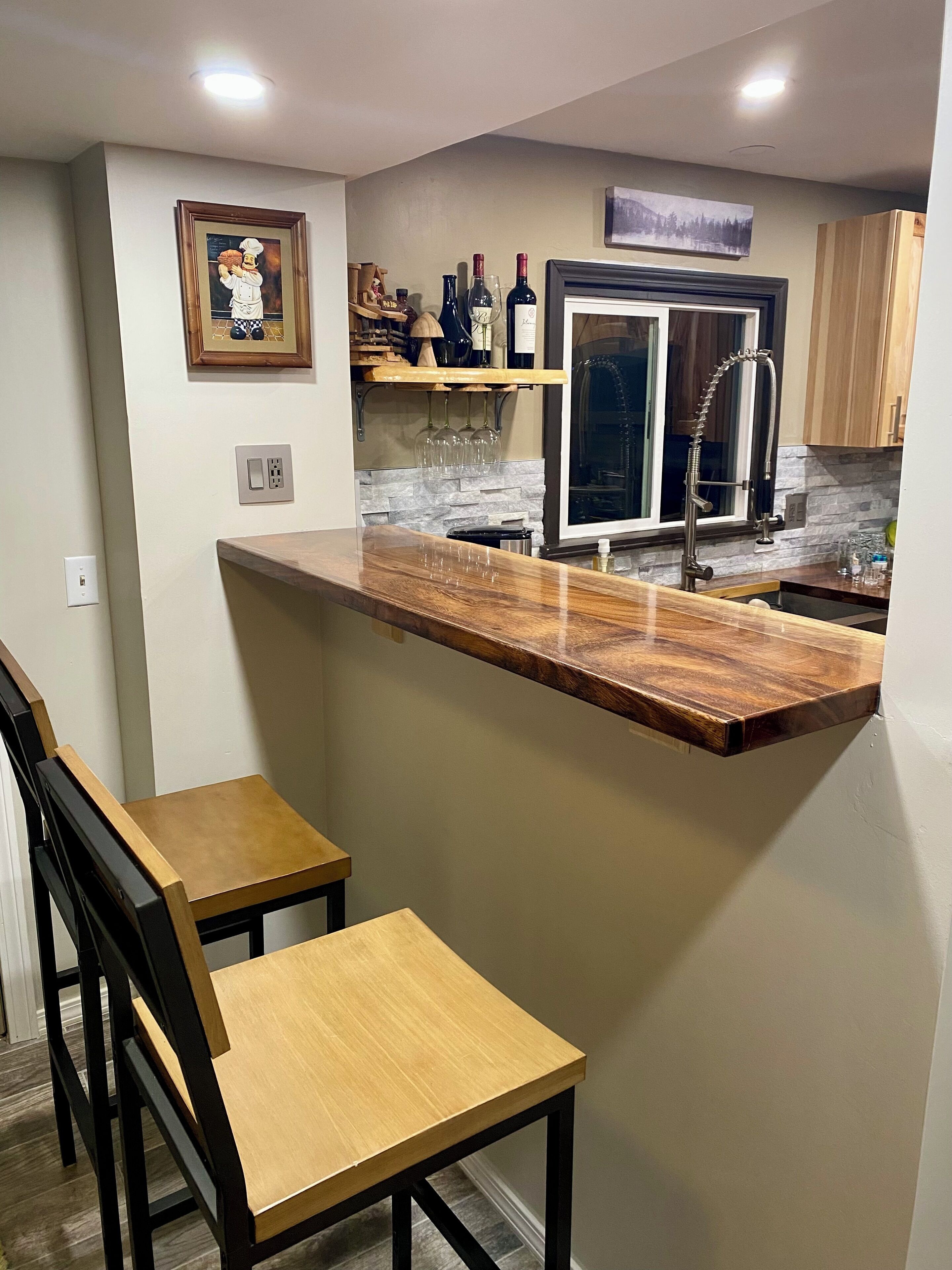 Une cuisine moderne dotée d’un comptoir-bar en bois, de deux tabourets de bar, d’un meuble à vin intégré et d’un réfrigérateur en acier inoxydable.