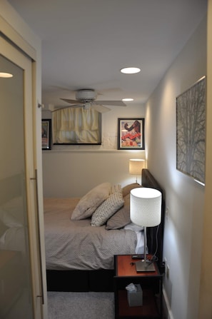 Ein Schlafzimmer mit einem Bett, einem Nachttisch mit Lampe, einem Deckenventilator und einem gerahmten Bild an der Wand.