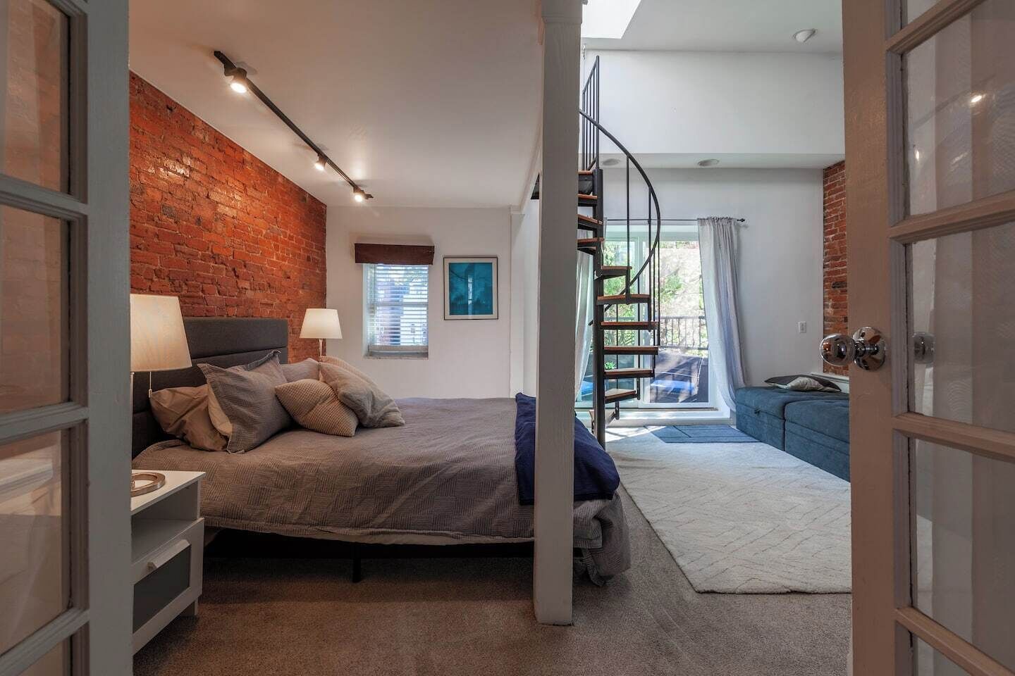 Een slaapkamer met een bakstenen muur, een bed met kussens, een nachtkastje, een raam met jaloezieën, een trap en uitzicht op buiten.