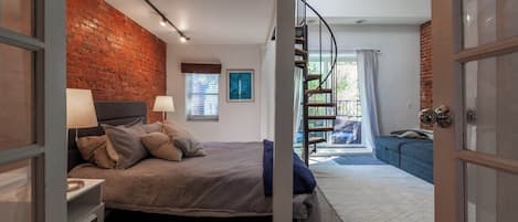 Quarto com parede de tijolos, cama com almofadas, mesa de cabeceira, janela com persianas, escada e vista para o exterior.