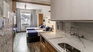 Ett kompakt hotellrum med ett litet pentry, en säng med blått sänglinne, en takfläkt och utsikt över omgivningarna.