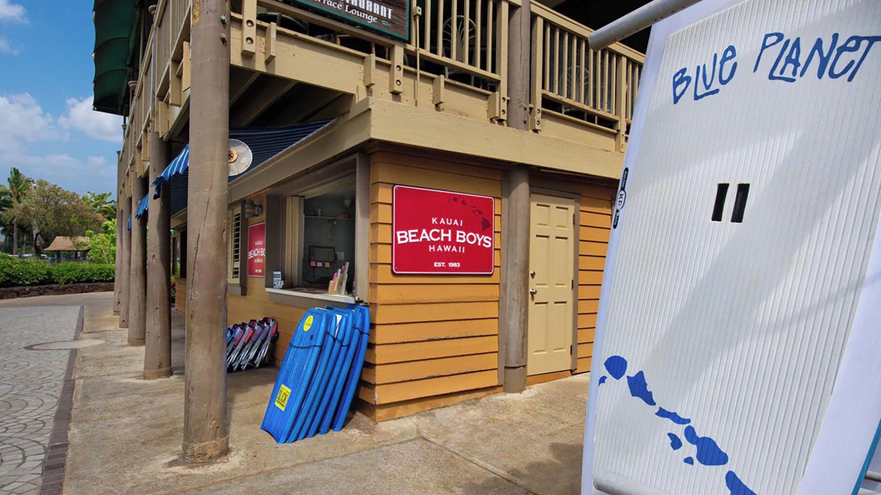 Ein restaurant im Strandhausstil mit einem Schild mit der Aufschrift "Kauai Beach Boys Hawaii" und einer blauen Surfbrett-Ausstellung draußen.