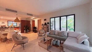 Ein modernes Wohnzimmer mit einer Couch, Sesseln und einem Couchtisch. Es gibt einen Essbereich mit Tisch und Stühlen, und im Hintergrund eine Küche.