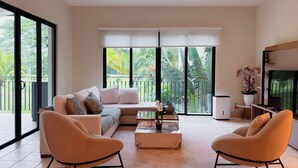 Ein modernes Wohnzimmer mit einer Couch, zwei Sesseln, einem Couchtisch und einem Fernseher. Große Fenster bieten Blick auf Grünfläche vor dem Haus.