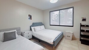 Ein Schlafzimmer mit zwei Betten, einem Nachttisch, einem Fenster mit Blick auf Bäume und einer Deckenleuchte.