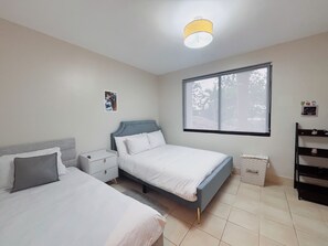 Et soveværelse med to senge, et natbord, et vindue med udsigt til træer og en loftlampe.