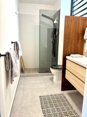 Un baño con cabina de ducha de vidrio, inodoro con asiento de madera, lavamanos con tocador de madera y una alfombra estampada en el suelo.