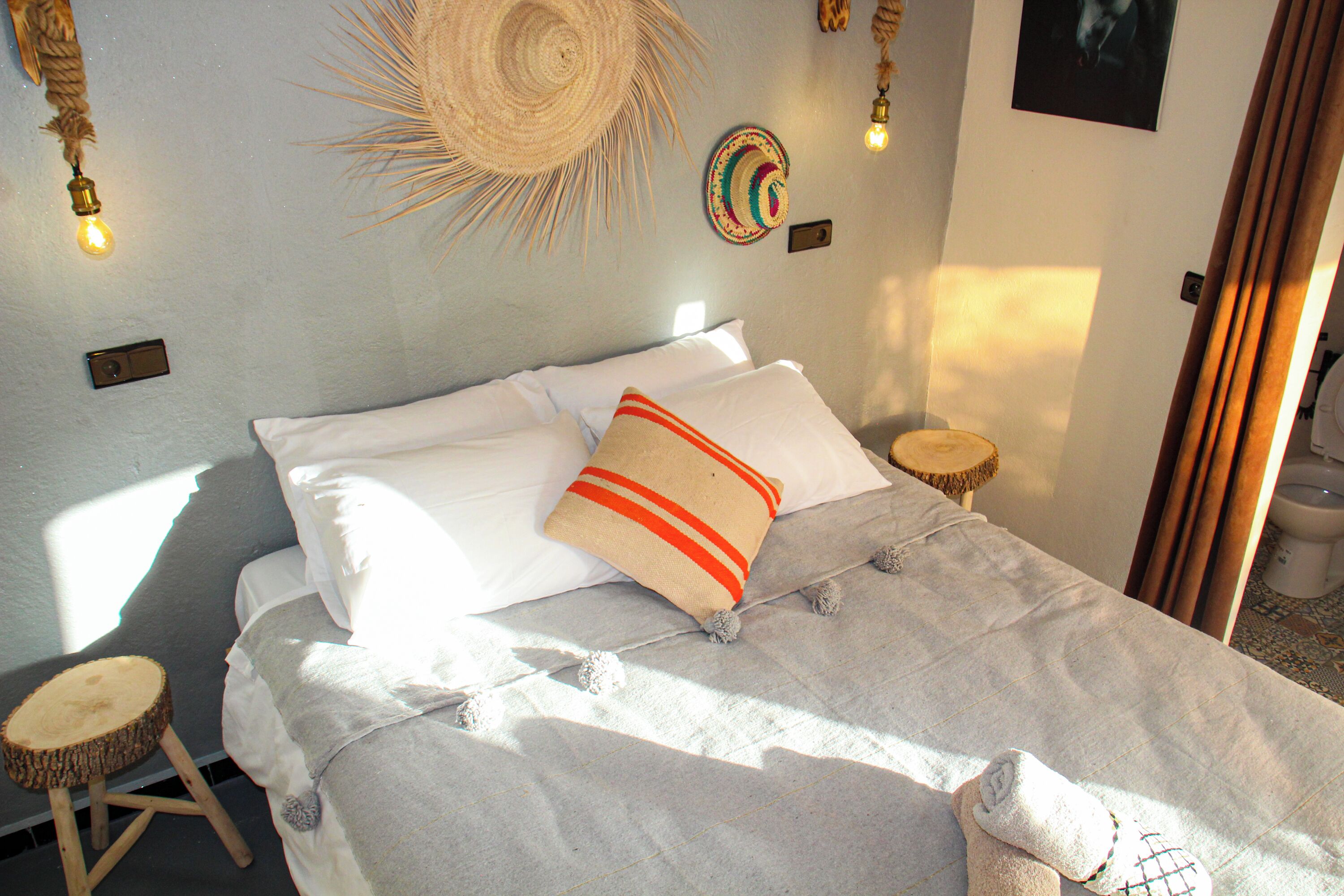 A bed with white linens, a beige pillow with red stripes, and a woven headboard. There are two bedside tables with woven bases, a wall-mounted light fixture, and a wall-mounted clock.