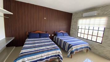 A room with two beds, wooden headboards, a window with blinds, and an air conditioning unit.