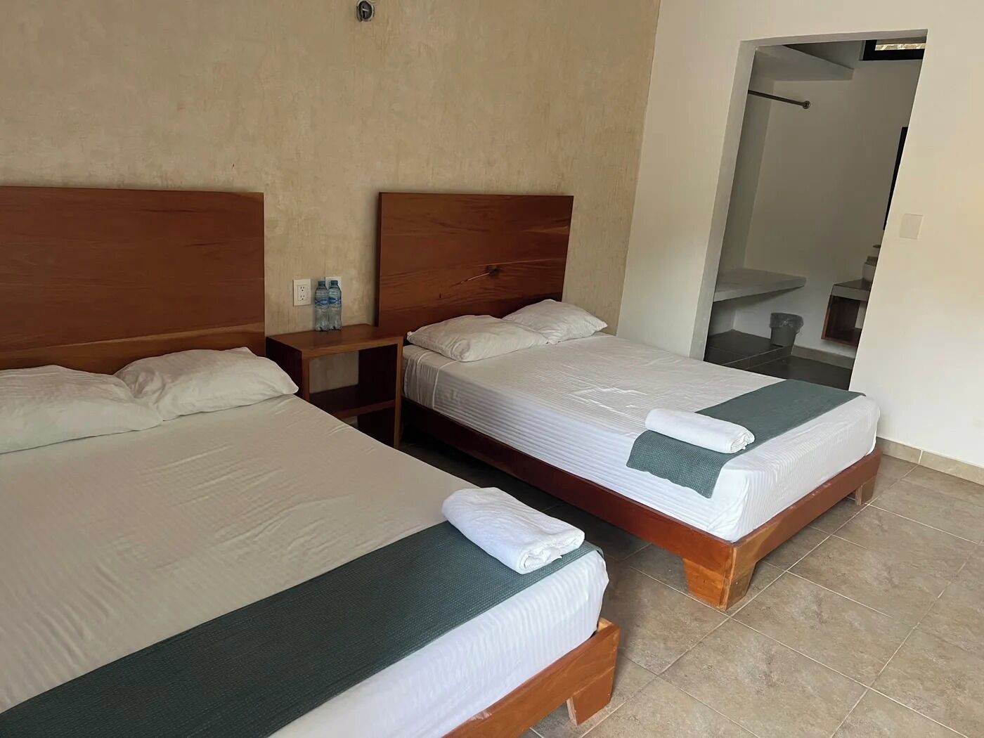 A hotel room with two single beds, a wooden headboard, a nightstand with a water bottle, and a mirror on the wall.