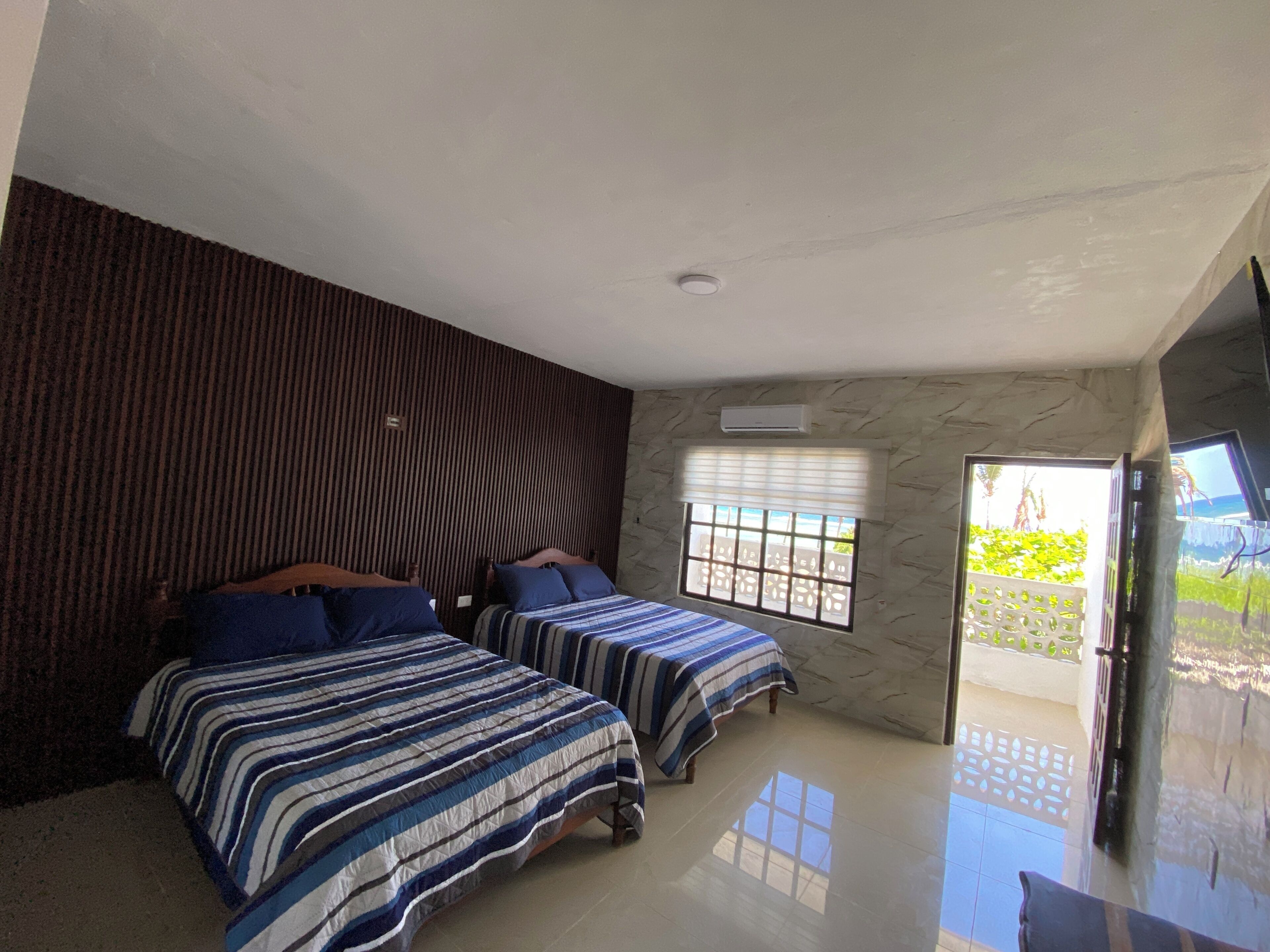 A bedroom with two beds, a window with blinds, a door leading outside, and a flat-screen TV mounted on the wall.