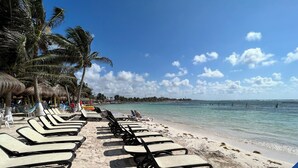 Una playa con filas de tumbonas y sombrillas de paja, un cielo azul despejado y un mar tranquilo.