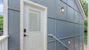 Een grijs gebouw met een witte deur, een klein buitenlampje en een metalen leuning.