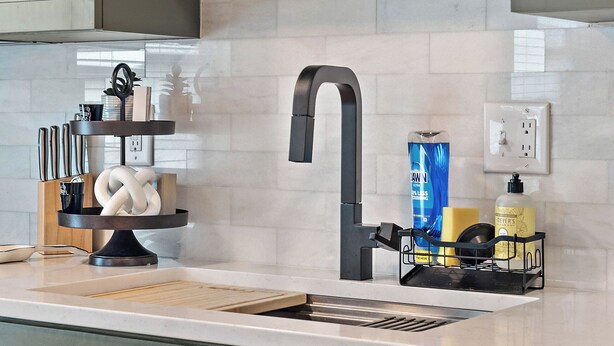 Een keuken met een modern design, met lichte kastjes, een roestvrijstalen spoelbak en een zwarte kraan. Er is een plank met keukengerei en een fles afwasmiddel.