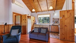 Interior acolhedor de uma cabana com teto de madeira, um sofá com almofadas estampadas, uma área de jantar com janela e uma cozinha com frigorífico e micro-ondas.