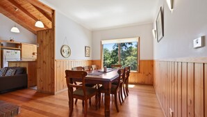 Uma sala de jantar acolhedora com mesa e cadeiras de madeira, uma cozinha com armários de madeira e uma grande janela com vista para o verde.