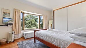 Quarto com cama, janela com cortinas, radiador, televisão em suporte e um quadro na parede.