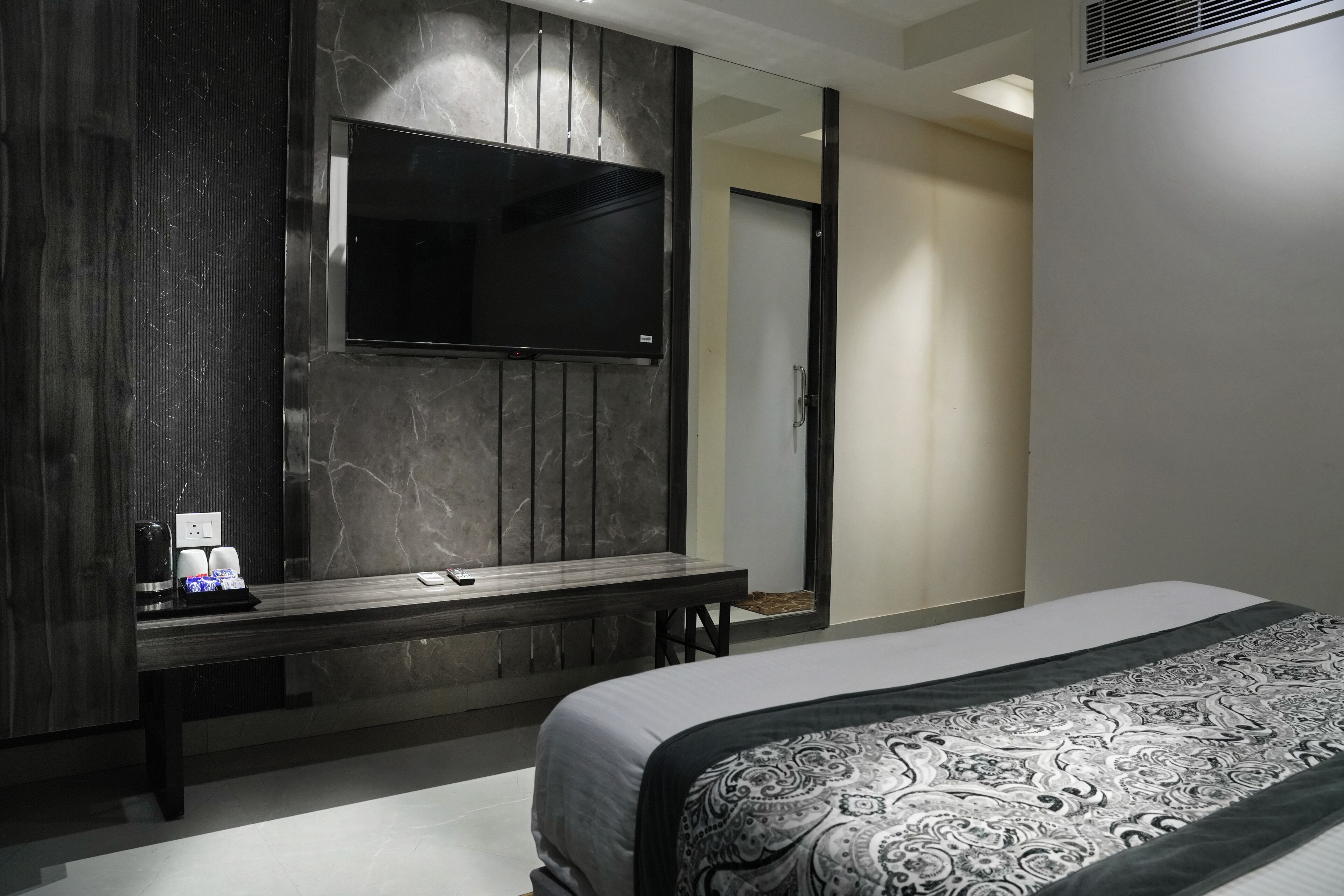 Una habitación de hotel moderna con una cama grande, una mesita de noche con cafetera, un televisor montado en un panel de madera oscura y una puerta que da a un baño.