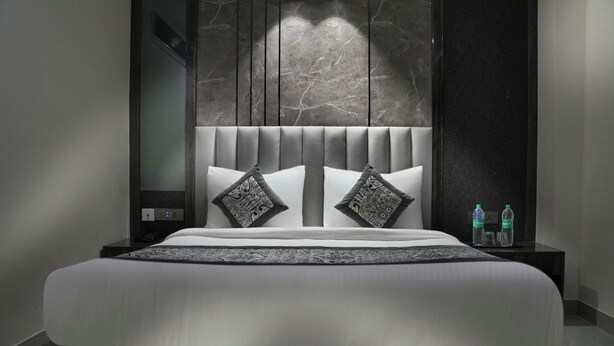 Une chambre d’hôtel moderne avec un grand lit, une tête de lit ornée de lignes verticales, deux coussins à motifs, une table de chevet avec des bouteilles d’eau et un mur à la texture évoquant le marbre.