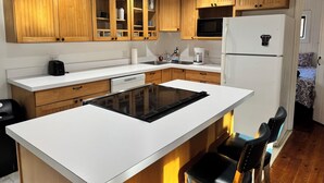 Una cocina moderna con armarios de madera, una placa de inducción negra, un refrigerador blanco y una zona de comedor con sillas negras.