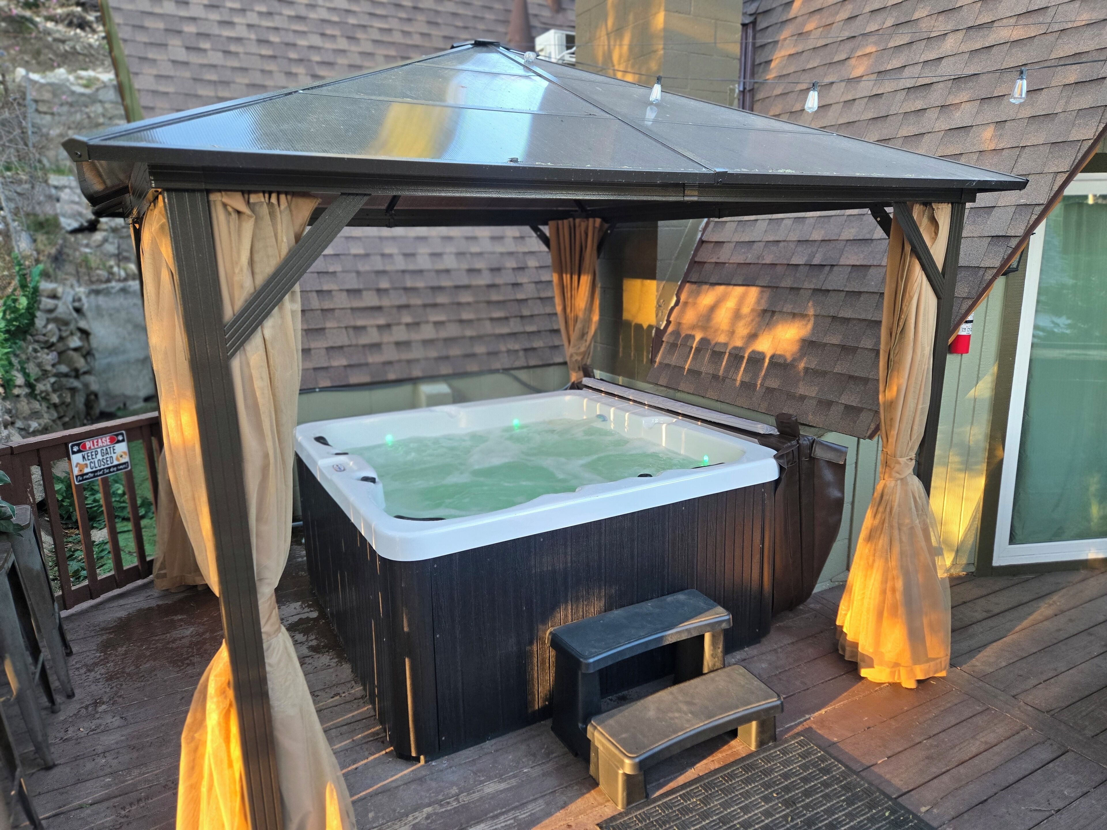 Ein Whirlpool unter einer Pergola mit einer Holterrasse und einem Schild mit der Aufschrift 'Bitte Tor geschlossen halten'.
