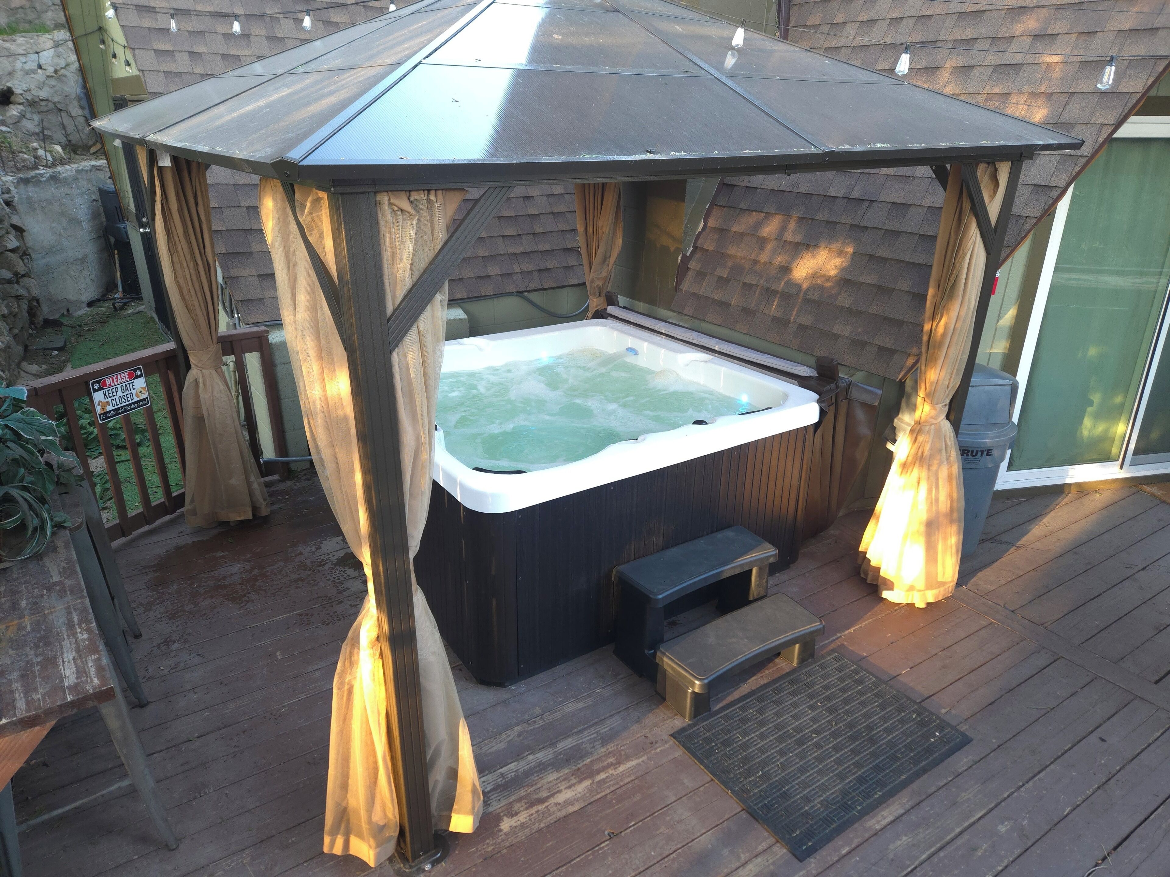 Un jacuzzi bajo un porche de madera con un banco y un basurero cerca.