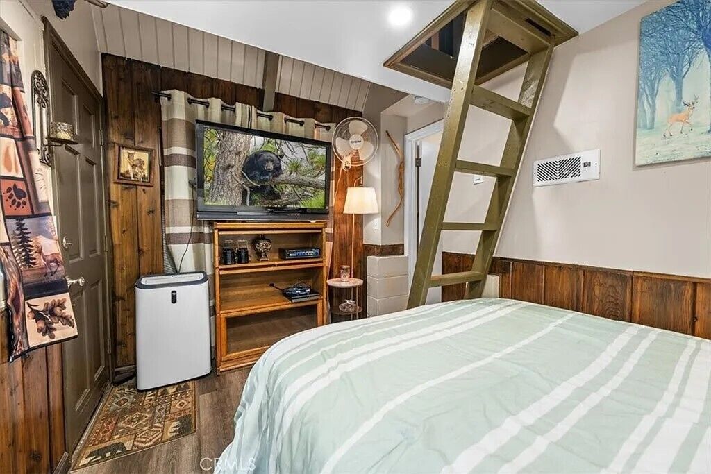A bedroom with a bed, a TV, a small fridge, a ladder, and a fan.