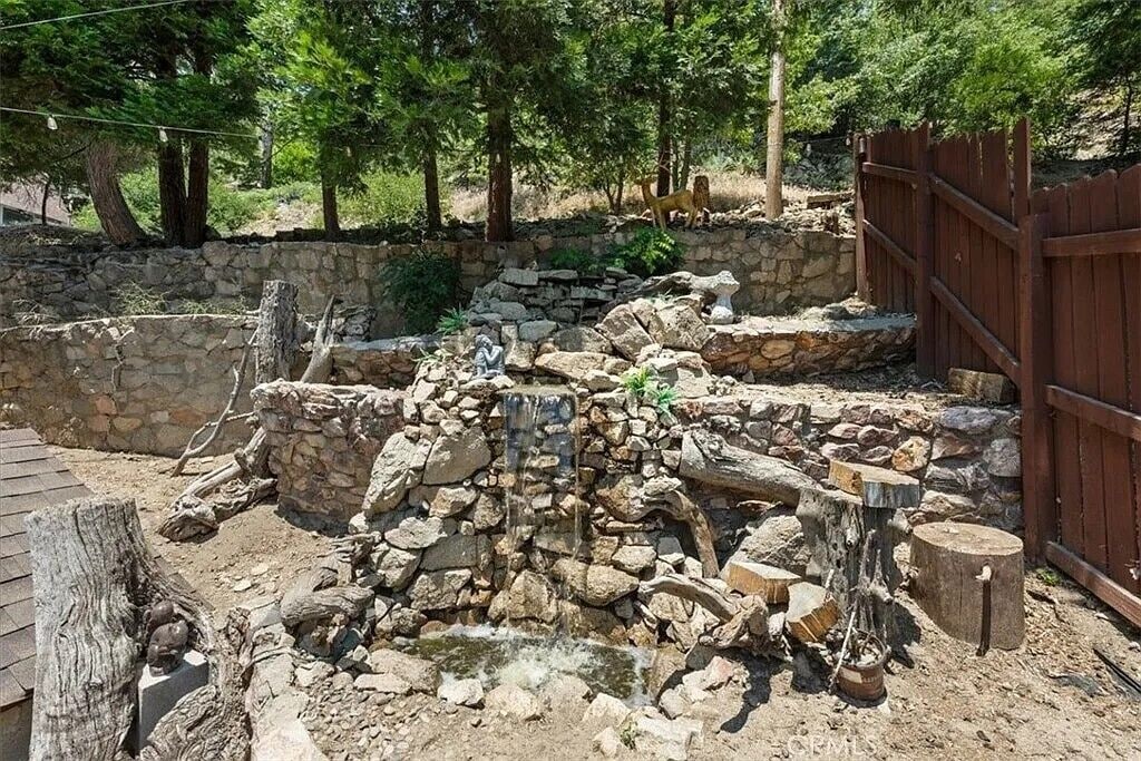 Een rustieke omgeving buiten, met een stenen gebouw, houten omheining en een kleine waterval.