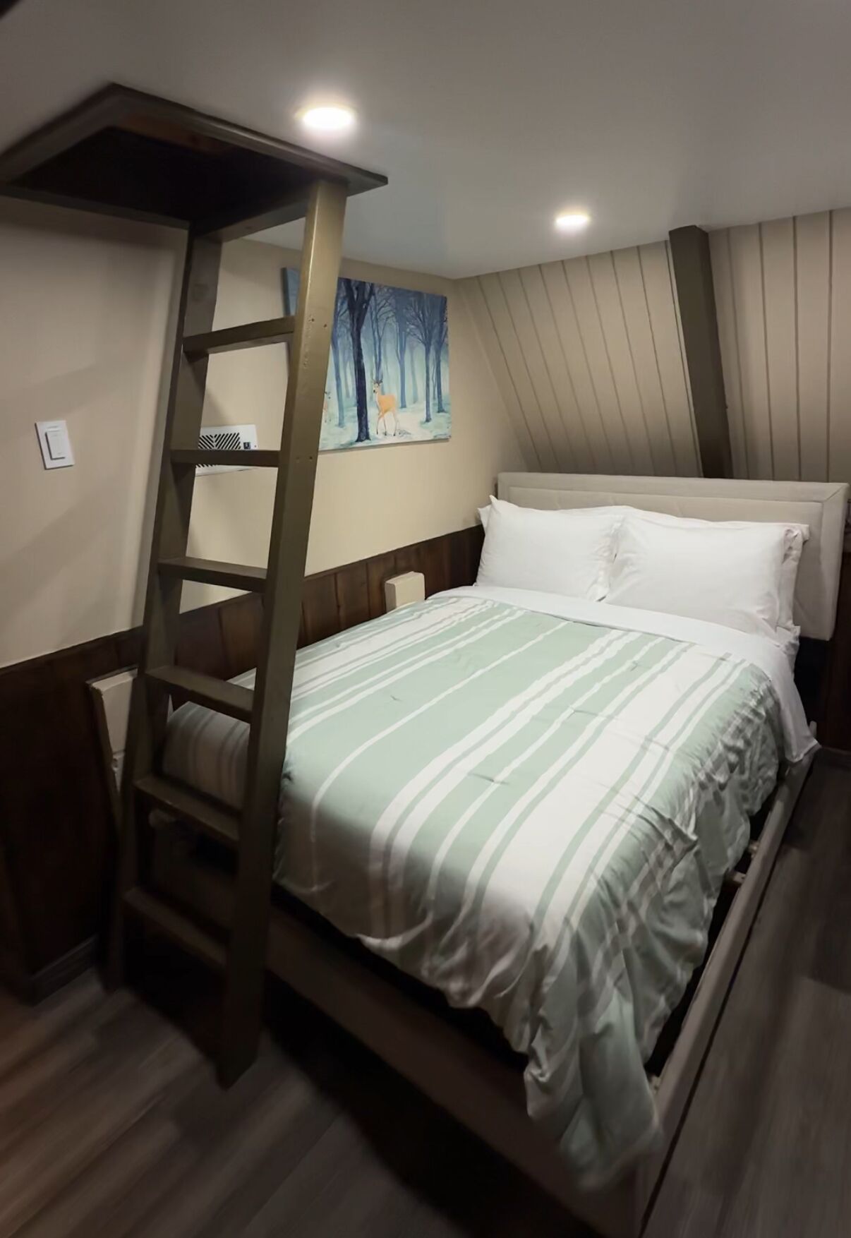 A bedroom with a bunk bed, a ladder, a painting of a forest scene, and a bed with white bedding.