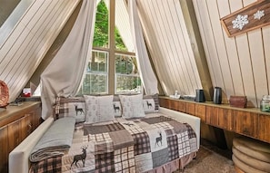 Ein gemütliches Schlafzimmer mit schräg stehender Decke, einem Bett mit Karo-Bettwäsche mit Rentier-Muster und einem Fenster mit Vorhängen.