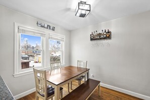 A dining area with a wooden table and chairs, large windows with blinds, and a wine rack on the wall.