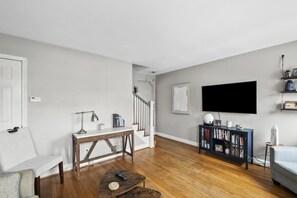 A living room with a wooden floor, a white sofa, a coffee table, a bookshelf, and a TV mounted on the wall.