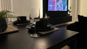 Ein modernes Wohnzimmer mit einem dunkelholzigen Couchtisch, einem Weinglas, einer schwarzen Schale und einem Topfpflanzen. An der Wand ist ein Flachbildfernseher angebracht, der eine farbenfrohe Szene zeigt.