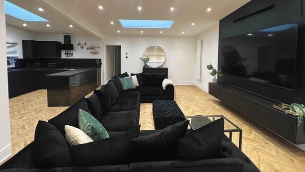 Ein modernes Wohnzimmer mit einem großen Flachbildfernseher, schwarzen Sofas und einer Küchenzeile im Hintergrund.
