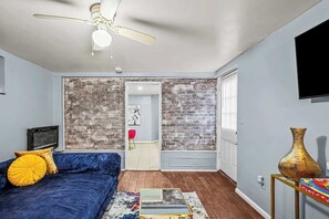 Un salon avec un canapé bleu, un ventilateur de plafond, un mur décoré d’un motif en briques, un téléviseur et un vase posé sur une table d’appoint.